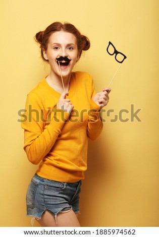 Cheerful young redhead woman dressed casually holding party props and posing over yellow background.