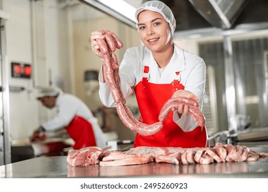 Cheerful young salesgirl of butcher shop in white uniform and red apron holding chain of raw pork sausages, offering fresh meat produce - Powered by Shutterstock