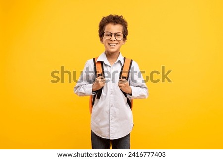Cheerful young schoolboy with curly hair and glasses, gripping his backpack straps, ready for new school day against yellow background