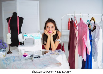 Cheerful young seamstress sits at the table with working things and own collection, in atelier