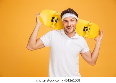 Cheerful young skater holding a skateboard over his shoulders and looking at the camera isolated on orange background