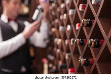 Cheerful young sommelier is choosing wine in cellar. He is smiling. Focus on bottles in shelf - Powered by Shutterstock