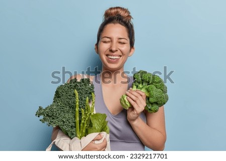 Cheerful young sportswoman with hair bun poses with fresh green vegetables keeps to healthy nutrition smiles happily keeps eyes closed isolated over blue background. Diet detox and sport concept
