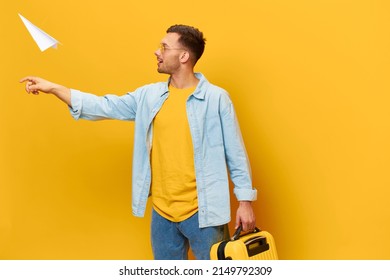 Cheerful Young Tanned Handsome Man In Blue Shirt Trendy Eyewear Throws Paper Airplane Hold Suitcase Posing Isolated On Orange Yellow Studio Background. Copy Space Banner Mockup. Trip Journeys Concept