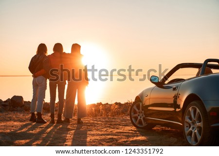 Cheerful young three women standing near cabriolet wathing at sunset