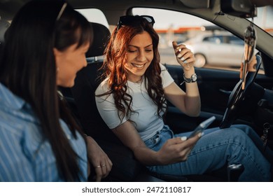 Cheerful young trendy women sitting in a car in front seats and using cellphone for GPS during their journey. Happy female friends having fun with mobile phone while sitting in a car during road trip. - Powered by Shutterstock