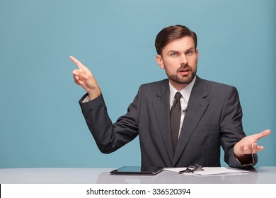 Cheerful Young Tv Newscaster Is Reporting News. He Is Sitting At The Desk And Looking At Camera With Surprise And Question. The Man Is Pointing Finger Sideways. Isolated And Copy Space In Left Side 