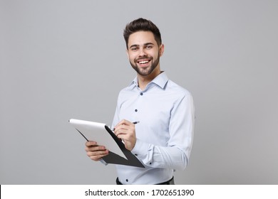 Cheerful young unshaven business man in light shirt posing isolated on grey background. Achievement career wealth business concept. Mock up copy space. Hold clipboard with papers document write notes - Shutterstock ID 1702651390