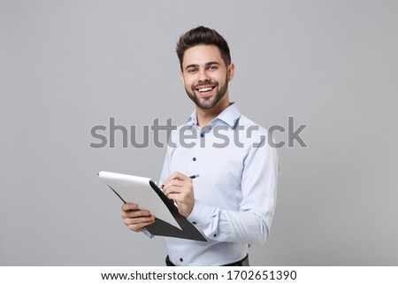 Cheerful young unshaven business man in light shirt posing isolated on grey background. Achievement career wealth business concept. Mock up copy space. Hold clipboard with papers document write notes