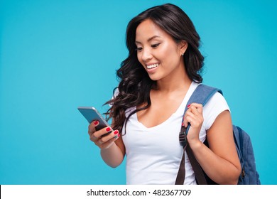Cheerful young vietnamese girl with backpack using smartphone isolated on a blue background