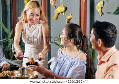 Cheerful young Vietnamse woman serving spicy soba noodle for daughter and husband at dinner