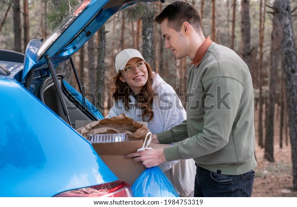 cheerful young volunteers holding carton box with\
rubbish near car 
