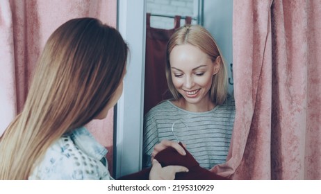 Cheerful Young Woman Is Appearing From Behind Curtain Of Fitting Room While Friendly Shop Assistant Is Giving Her New Jumper To Try On. Customer Is Taking Garment And Drawing Curtain Back.