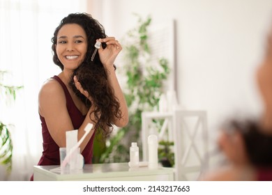 Cheerful Young Woman Applying Repair Serum On Damaged Hair Near Mirror At Home, Empty Space. Lovely Millennial Lady Using Organic Cosmetics For Haircare, Enjoying Daily Beauty Routine