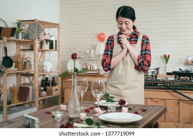 cheerful young woman in apron standing at set up romantic kitchen table with flowers and roses smiling waiting for husband coming back celebrating valentine day dinner. decorated dining room concept - Shutterstock ID 1377719099