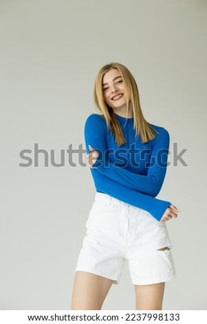 cheerful young woman in blue zipped turtleneck and white shorts posing isolated on grey