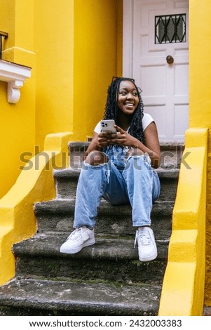 A cheerful young woman with braided hair sits on yellow stairs while texting on her smartphone, embodying a moment of modern connectivity and urban style.