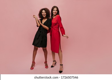 Cheerful young woman with brunette hair in red dress and black shoes smiling and posing with curly friend in dark clothes.
