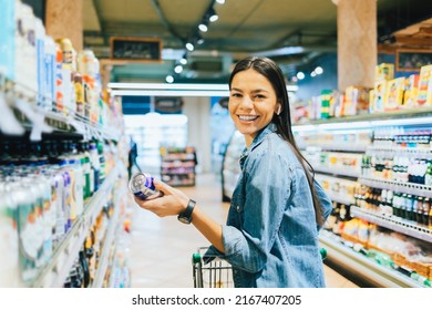 Cheerful Young Woman Buying Beer Or Ale Alcohol In Liquor Store, Standing In Front Of Shelf Holding Can Smiles Looking At Camera