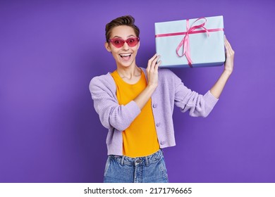 Cheerful Young Woman Carrying Gift Box On Shoulder While Standing Against Purple Background