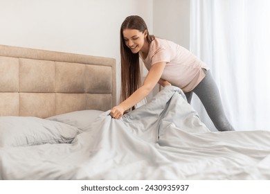 Cheerful young woman in casual attire smiling while tidying up and making her bed in bright, airy bedroom, indicating fresh start to her day, copy space