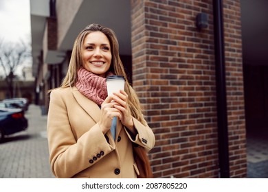 A Cheerful Young Woman In A Coat And With A Scarf Is Holding Hot Coffee In Her Hands. On Cold Days, We Have A Hot Beverage To Warm Us. A Woman With A Coffee Standing Outside.