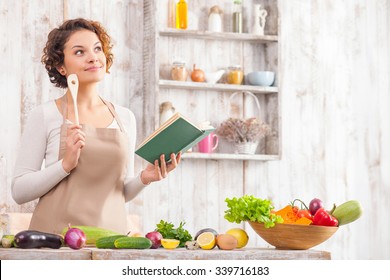 Cheerful young woman is cooking in the kitchen with joy. She is standing and holding a book of recipe. The lady is touching a wood spoon to her face and dreaming. She is smiling