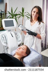 Cheerful young woman cosmetologist holding medical ultrasound device and smiling while standing by client. Charming female patient lying on daybed while having SMAS lifting procedure in modern clinic.