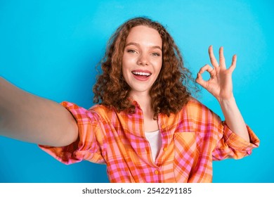 Cheerful young woman with curly hair gives an okay gesture in a vibrant plaid shirt against a blue background, expressing joy and positivity in a casual setting. - Powered by Shutterstock