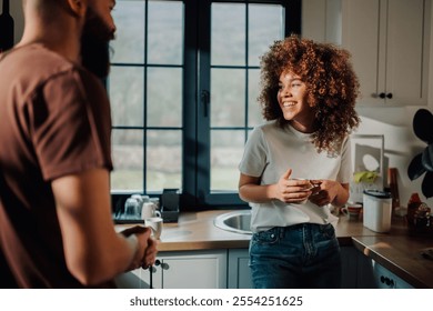 Cheerful young woman with curly hair smiling at her partner while enjoying a cup of coffee in a sunlit kitchen, embodying a cozy domestic moment - Powered by Shutterstock