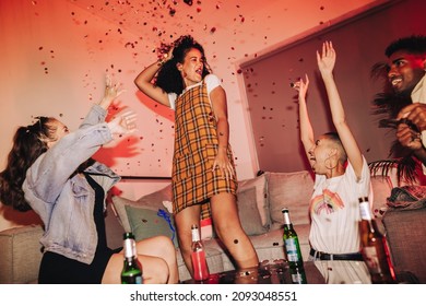 Cheerful young woman dancing with her friends under falling confetti. Group of happy friends enjoying their favourite song at a house party. Multicultural friends having fun together on the weekend.