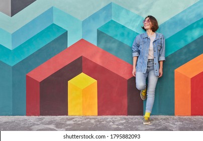 Cheerful young woman in denim clothes smiling and looking away while leaning on colorful wall with geometric graffiti in city - Shutterstock ID 1795882093