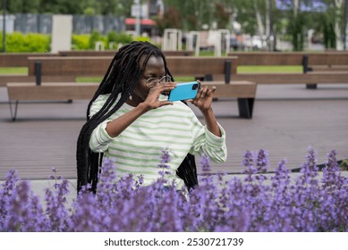 Cheerful young woman with dreadlocks joyfully captures a picture of vibrant lavender blossoms in a sunny park with her smartphone - Powered by Shutterstock