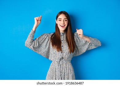 Cheerful Young Woman In Dress Raising Hands Up And Smiling Excited, Triumphing Over Winning Prize, Celebrating Victory, Standing On Blue Background