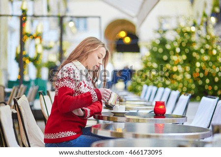 Cheerful young woman drinking hot chocolate in a cozy outdoor Parisian cafe decorated for Christmas