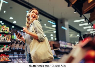 Cheerful young woman with earphones listening music, having fun while buying some groceries in supermarket