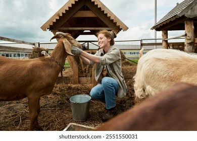 Cheerful young woman feeding purebred goat and petting animal while sitting on squats in the yard of livestock farm - Powered by Shutterstock