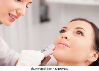 Cheerful young woman is getting botox procedure. The doctor is standing near her and smiling. She is holding syringe and touching it to female face
