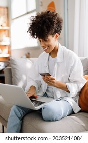Cheerful young woman going on an online shopping spree. Young woman smiling while using her credit card to make a purchase on her laptop. Woman sitting alone on a couch at home.