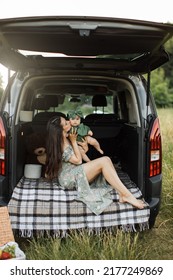 Cheerful Young Woman In Green Summer Dress Lifting Up Six Month Old Baby Girl While Playing Sitting On Car Trunk Outdoors. Mother With Daughter Enjoying Time Spending Together On Fresh Air.