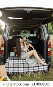 Cheerful Young Woman In Green Summer Dress Lifting Up Six Month Old Baby Girl While Playing Sitting On Car Trunk Outdoors. Mother With Daughter Enjoying Time Spending Together On Fresh Air.