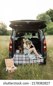 Cheerful Young Woman In Green Summer Dress Lifting Up Six Month Old Baby Girl While Playing Sitting On Car Trunk Outdoors. Mother With Daughter Enjoying Time Spending Together On Fresh Air.