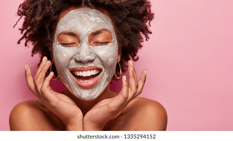 Cheerful young woman has Afro hairstyle, touches cheeks, has clay mask on face, enjoys softness, has beauty treatments at home, poses half naked, isolated over pink background. Skin care concept