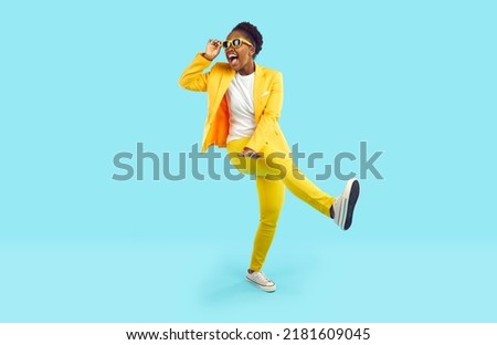 Cheerful young woman having fun, dancing and laughing isolated on light blue background. African American young woman in yellow suit and sunglasses is walking with funny expression on her face.