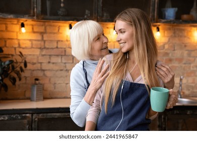 Cheerful young woman having rest and drinking tea with her senior mother while listening music in headphones and smiling to each other. Happy family concept. Two generations of woman in kitchen - Powered by Shutterstock