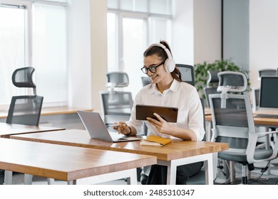 A cheerful young woman with headphones works on a tablet and laptop in a bright, modern office setting.

 - Powered by Shutterstock