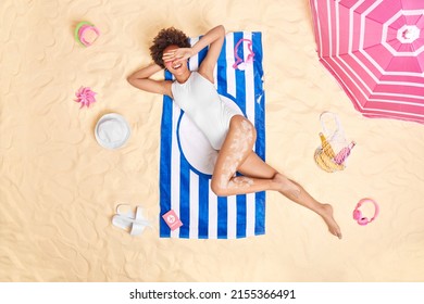 Cheerful young woman hides face with hand smiles gladfully wears white swimsuit poses on striped towel sunbathes on beach has lazy day travels abroad for summer vacation. Holidays time concept