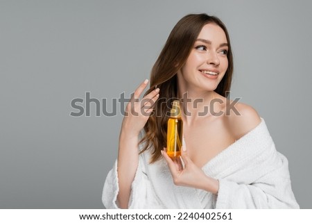 cheerful young woman holding bottle with hair oil isolated on grey