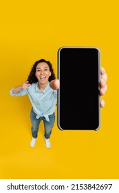 Cheerful young woman holding cell phone in hand pointing at gadget with blank black mockup screen smiling on yellow orange wall, recommending newest mobile app, above high angle top view, copy space - Shutterstock ID 2153842697
