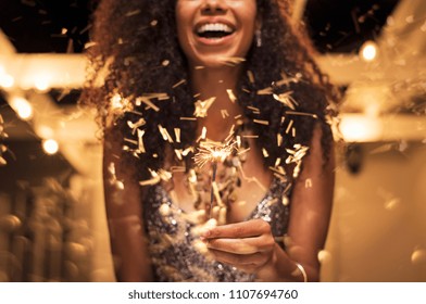 Cheerful young woman holding single sparkler in hand outdoor. Detail of african girl celebrating new year’s eve with bengal light. Closeup of beautiful woman holding a sparkling stick at party night. - Powered by Shutterstock